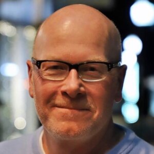 Smiling bald man with black rectangular glasses and light stubble, indoors with blurred lights in the background